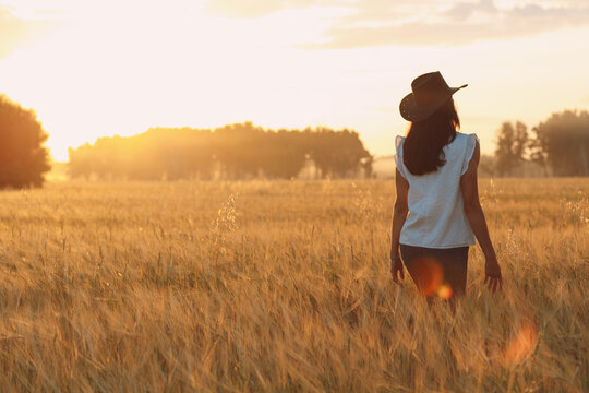 Woman farmer in cowboy hat walking with hands on ears at agricultural barley field on sunset.