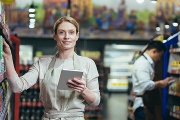 Portrait of an experienced female seller in a supermarket, the manager is looking at the camera and smiling, holding a tablet computer in her hands, the saleswoman is among the shelves with goods in