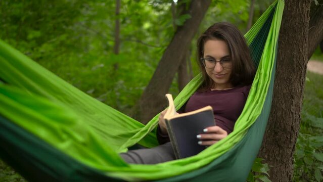 A young woman lies in a hammock and reads a book. A girl with glasses in nature is engaged in self-development.