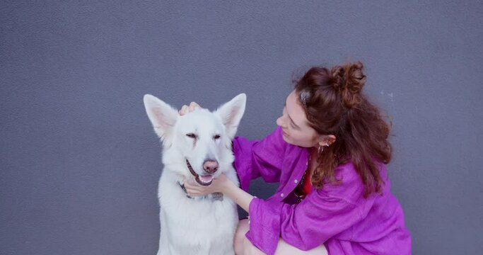a cute girl with curly hair hugs and plays with a white swiss shepherd, the dog looks in the eyes and sticks out its pink tongue. casual girl talking with a dog near the wall. middle plan
