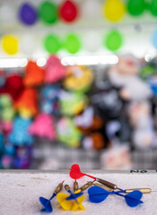 selective focus on darts sitting on a ledge with a wall of balloon targets and prizes that a player can win at a fair.