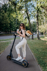 Happy smiling woman traveler is standing with electro scooter and drinking coffee in city parkland with dog Welsh Corgi Pembroke in a special backpack