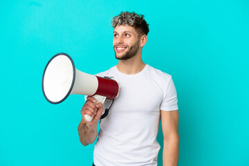 Young handsome caucasian man isolated on blue background holding a megaphone and looking up while smiling