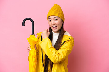 Young Chinese woman with rainproof coat and umbrella isolated on pink background celebrating a...