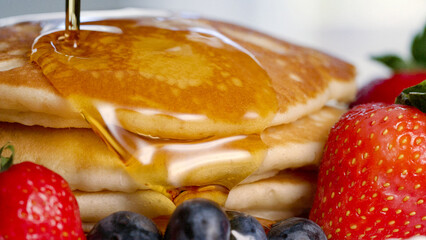 Close shot of maple syrup poured on stacked pancakes with berries around