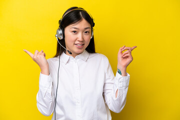 Telemarketer Chinese woman working with a headset isolated on yellow background pointing finger to the laterals and happy