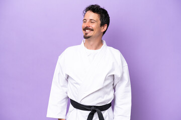 Young caucasian man doing karate isolated on purple background looking to the side and smiling