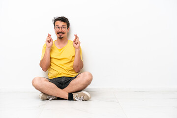 Young caucasian man sitting on the floor isolated on white background with fingers crossing