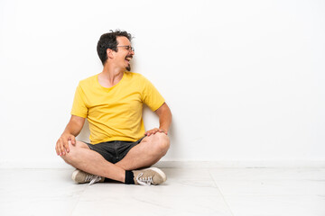 Young caucasian man sitting on the floor isolated on white background laughing in lateral position