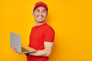 Smiling young Asian man in red cap t-shirt uniform, employee work as dealer courier, using laptop pc computer on work, look aside isolated on yellow background. Professional Delivery service concept