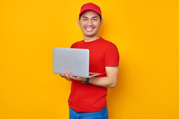 Smiling young Asian man in red cap t-shirt uniform, employee work as dealer courier, using laptop pc computer on work isolated on yellow background. Professional Delivery service concept