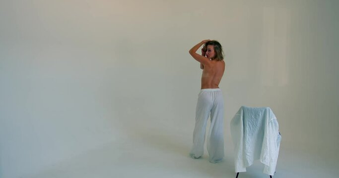 a girl stands on a white background in white pants and a bare back near a chair on which a white jacket. the girl holds on to her hair and turns to the camera. overall plan