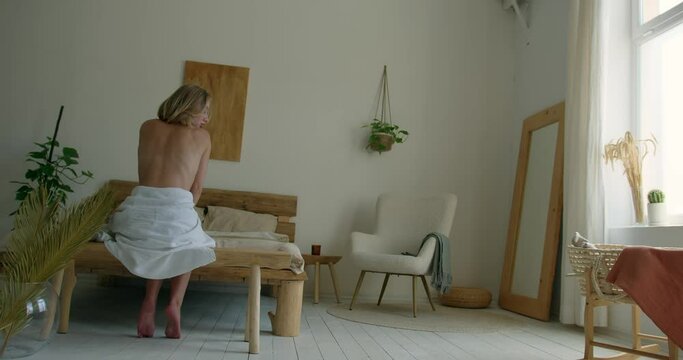 sexy blonde girl with a bare back sits on a wooden bench in the bedroom by the bed with her back to the camera and leans on her hand against the background of a large mirror chair window