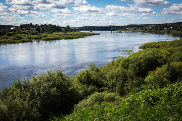 Russia Vologda Oblast Totma 08.12.2017 Summer  landscape: coastline the Sukhona River with blue sky and clouds reflecting in calm water, the village, green trees and grass, and the bridge far away