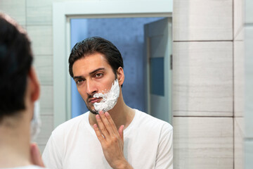 Handsome man looking at the mirror and shaving in the bathroom