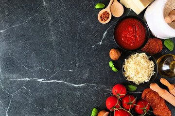 Pizza making ingredients including cheese, pepperoni, tomatoes and basil. Top view side border against a dark background. Copy space.