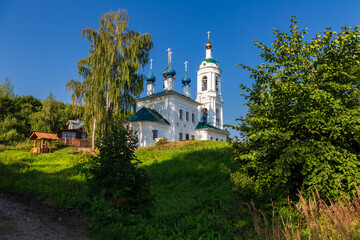 Old Russian Orthodox church under big birch tree on hill top against clear blue sky, Plyos, Russia