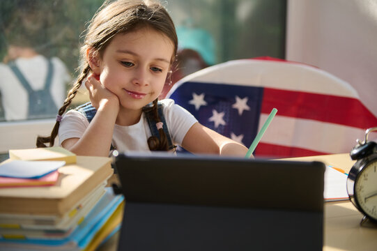 Charming little European girl, schoolgirl, first grader in an online school watching a video lesson on a digital tablet while studying remotely from home interior. Education across the distances