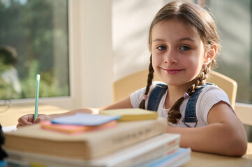 Fototapeta na wymiar Adorable Caucasian child, schoolgirl cute smiling looking at camera, sitting at a desk with folded textbooks on blurred foreground. Portrait of a happy girl first grader studying in an online school