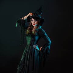 A charming young witch in a costume invites you to a sabbath, a Halloween party with costumes. Young brunette in pointed hat and black dress, photo on black