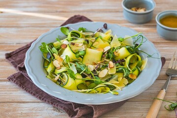 Healthy salad of raw zucchini, cheese, mix of microgreens and roasted almond slices with mustard dressing on a gray ceramic plate on a wooden background. Salad recipes.