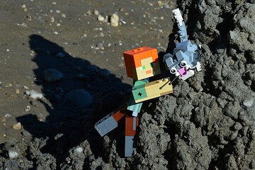 Fototapeta premium LEGO Minecraft large action figure of Alex character is helping LEGO cat down from tall black sand hill on the summer beach, daylight sunshine.