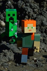 Fototapeta premium LEGO Minecraft figure of Alex chased by dangerous green explosive mob creature Creeeper on black sand muddy soil, summer daylight sunshine. 