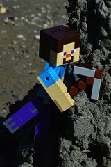 Fototapeta premium LEGO Minecraft large action figure of Steve with stone pickaxe, ready to mine some wet black sand from steep hill, summer daylight sunshine. 