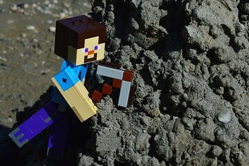 Fototapeta premium LEGO MInecraft large figure of Steve with stone pickaxe is mining wet black sand mud on hill on a beach, summer daylight sunshine. 