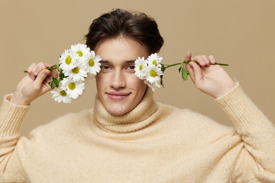 A gorgeousman with shining skin, with dark, short, short hair combed back, in a beige turtleneck with a high collar stands on a dark beige background with white daisies near his face.Close-up photo.