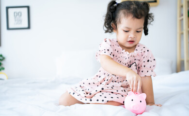 baby girl inserting a coin into piggybank. asian cute girl saving money putting coins into piggy bank.
kid saving money for future concept.