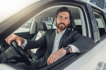 Handsome businessman in grey suit is riding behind steering wheel of car