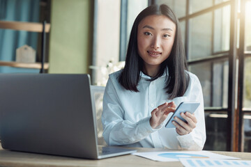 Asian business woman using phone while sitting in office and looking camera