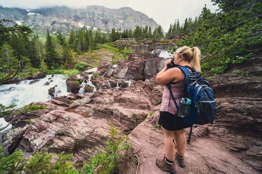 Blonde hiker woman takes photos at Redrock Falls along Swiftcurrent Pass hiking trail in Glacier National Park USA