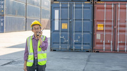 Asian elderly supervisor is using walkie talkie to communicate with his workers in a shipyard with cargo containers as background. Man in safety suit and helmet in a logistic place