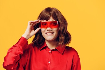 a stylish woman stands on a yellow background in a red shirt and glasses of an interesting shape, smiling pleasantly at the camera and adjusting her glasses