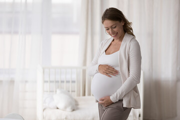 Young 30s happy pregnant woman in casual clothes embracing caressing her big belly, standing pose in light nursery at modern cozy home in last stages of pregnancy. Expectant mother, motherhood concept