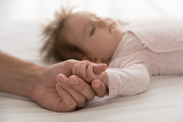 Loving daddy holds tiny arm cute sleeping newborn daughter, baby lying on bed touch fathers finger,...