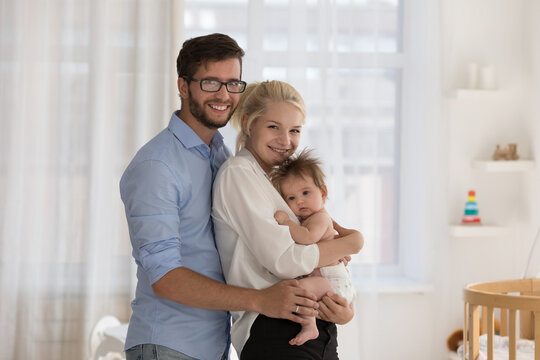 Portrait of young 30s loving parents, attractive cheerful Caucasian mother and father hugging their cute newborn baby boy or girl, standing in modern warm nursery, smile look at camera. Family concept