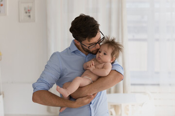 Young loving daddy standing in cozy warm domestic room hugs and kisses gently with love his cute newborn, lulls before daytime nap looking happy being father. Protection, adoption, family bond concept