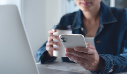 Young smart asian business woman sitting at table, drinking coffee and using mobile phone with laptop computer on table, have a coffee break during working at home office