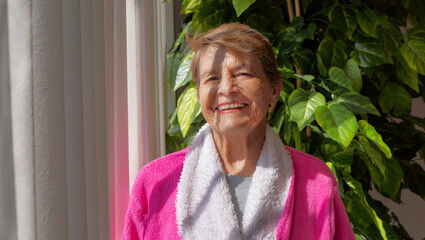 Older Mexican Latina woman smiling near a window in her bathrobe
