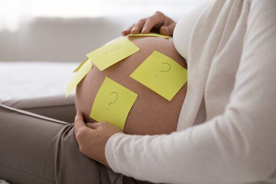 Close up crop shot, attached yellow sticky notes with questions marks on big bare belly of pregnant woman, touching caressing her abdomen. Think of name for future baby, guess gender of unborn concept