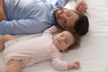 Obraz na płótnie Canvas Tired young father falls asleep near newborn baby girl, close up above shot. Sweet infant daughter sleep in bed with loving daddy enjoy daytime nap at home. Fatherhood, family, infancy, rest concept