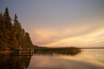 A boat dock by a summer cabin among reeds reflects in the water at sunset in Candle Lake, Northern...