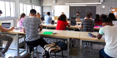 Rear view of group of multiracial high school students in class using laptops while teacher marks exams.