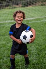 Obraz premium Young African American soccer player standing in front of a soccer goal smiling before a soccer game on a large grass field holding a ball