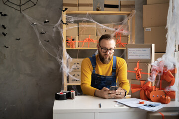 Small business owner man using mobile app on smartphone while checking parcel. Warehouse worker, seller in eyeglasses holding a phone. Big sale discounts and promotions of Halloween.