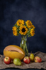 A bouquet of sunflowers in a round clay vase on a blue background. The fruits of autumn lie on craft paper: apples, pears, pumpkin. onions
