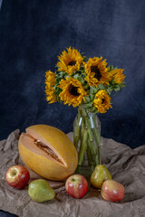 A bouquet of sunflowers in a round clay vase on a blue background. The fruits of autumn lie on craft paper: apples, pears, pumpkin. onions
